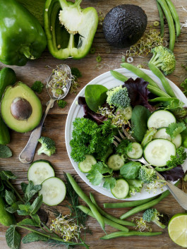 Chopped green vegetables and herbs in a plate and on the table: cucumbers, spinach, avocado, green peppers, peas, broccoli, mint, wheat sprouts, parsley