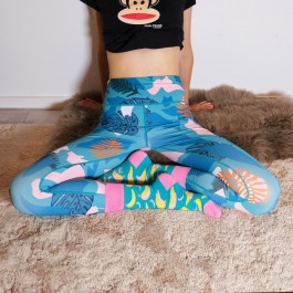 woman in black crop top, blue and pink socks with bananas and blue leggings with hawaiian multicolored print sits cross-legged on furs