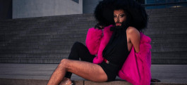 The drag queen poses seated in a black bodysuit, a pink short fur coat and a wide black fur hat 